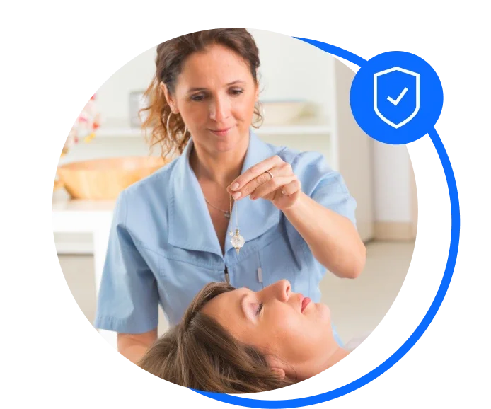 Female Complementary Therapist in a light blue blouse standing over a patient who is lying down with her eyes closed. The therapist is performing therapy.